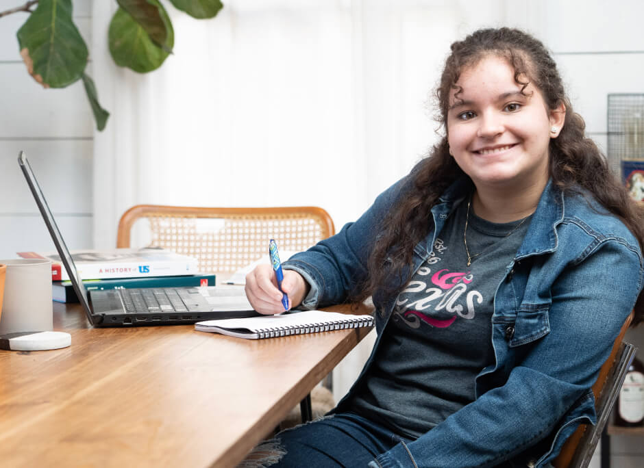 طالبة تبتسم للكاميرا وهي جالسة على طاولتها ومعها صورة الكمبيوتر المحمول الخاص بها